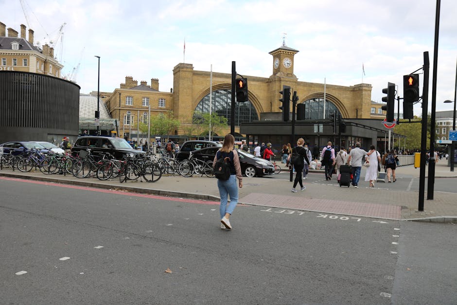 A busy urban scene outside a historic station entrance featuring a large, arched glass facade with a clock tower on top. In the foreground, a woman with light brown hair, wearing a beige jacket and li